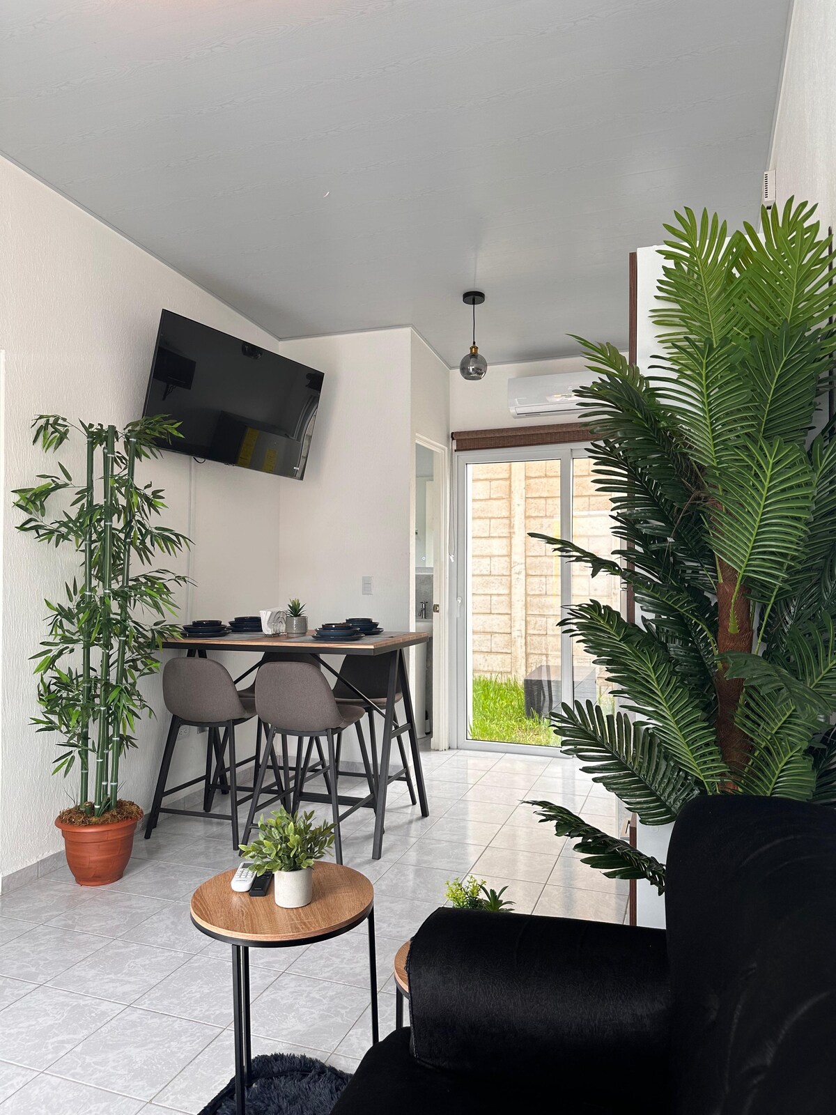 A modern dining area is visible with a black table set for four, complemented by stylish gray chairs. A large television is mounted on the wall, and indoor plants add a touch of greenery. Large windows provide natural light, leading to an outdoor space.
