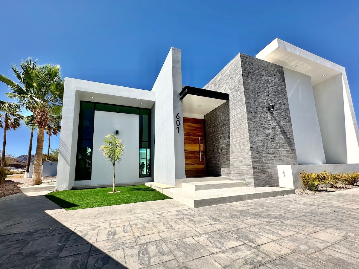 The modern exterior of the home is showcased with sleek lines and a combination of textured stone and smooth white walls. Lush palm trees and ornamental grasses are visible, framing a neat entrance with a wooden door and a small accent tree on the manicured lawn.