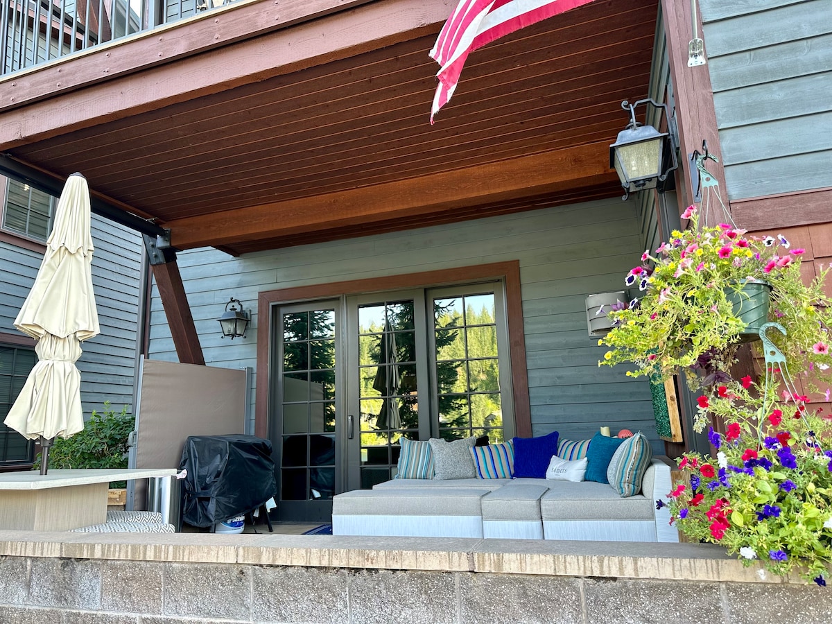 A covered patio area features a spacious sectional sofa adorned with colorful cushions and is complemented by hanging planters filled with vibrant flowers. An umbrella and grill are also visible, enhancing the outdoor living space beneath the upper deck.