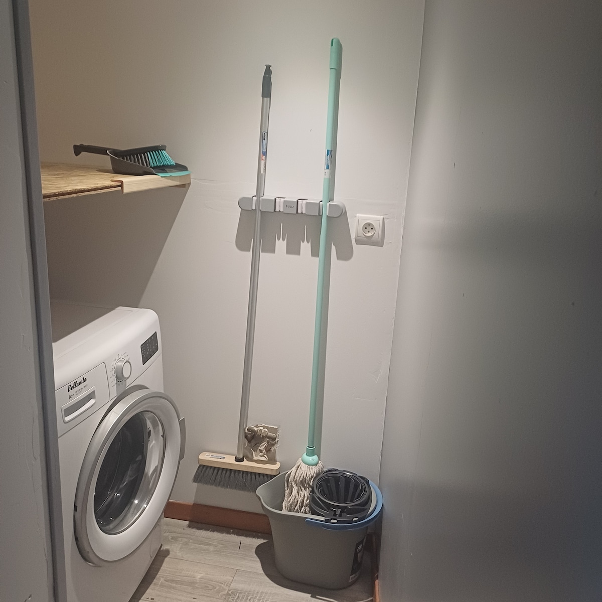 A cleaning area is equipped with a washing machine and a simple wooden shelf. Cleaning supplies are organized, including a broom, a mop, and a dustpan. The space features a neutral color palette and a vinyl floor, contributing to a functional atmosphere.