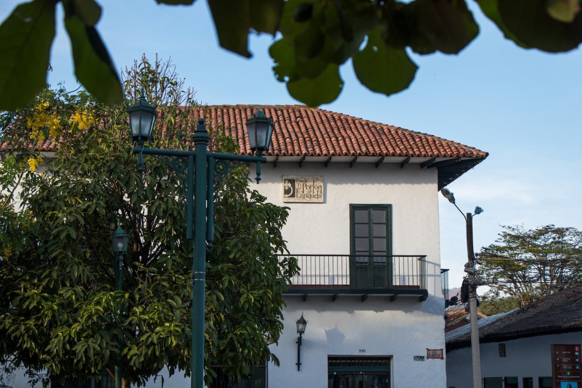 A traditional colonial house is showcased, featuring a sloped clay tile roof and a whitewashed facade. Green wooden shutters frame the windows, complemented by a balcony adorned with a decorative wooden railing. Lush trees provide shade in the foreground, enhancing the charming atmosphere.