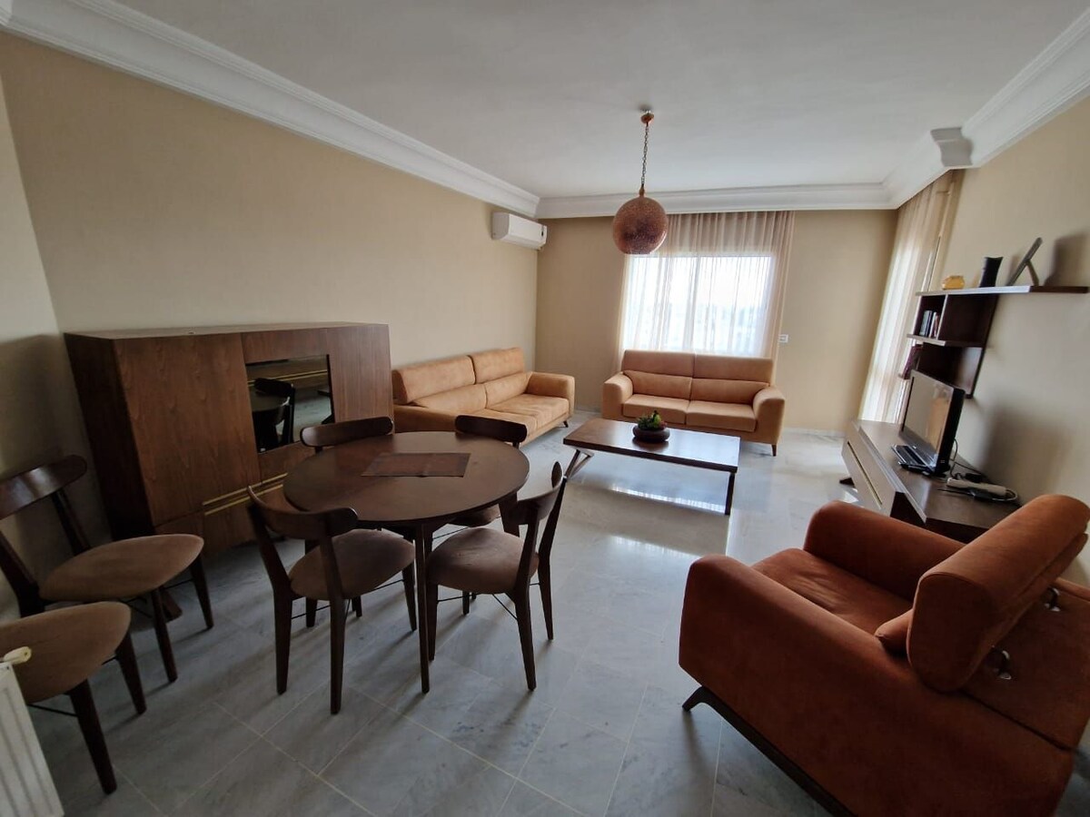 A spacious living area features two comfortable sofas and an elegant coffee table. Natural light enters through large windows, highlighting the neutral-colored walls and flooring. A dining table with six chairs provides ample seating, while a modern entertainment unit holds a television.