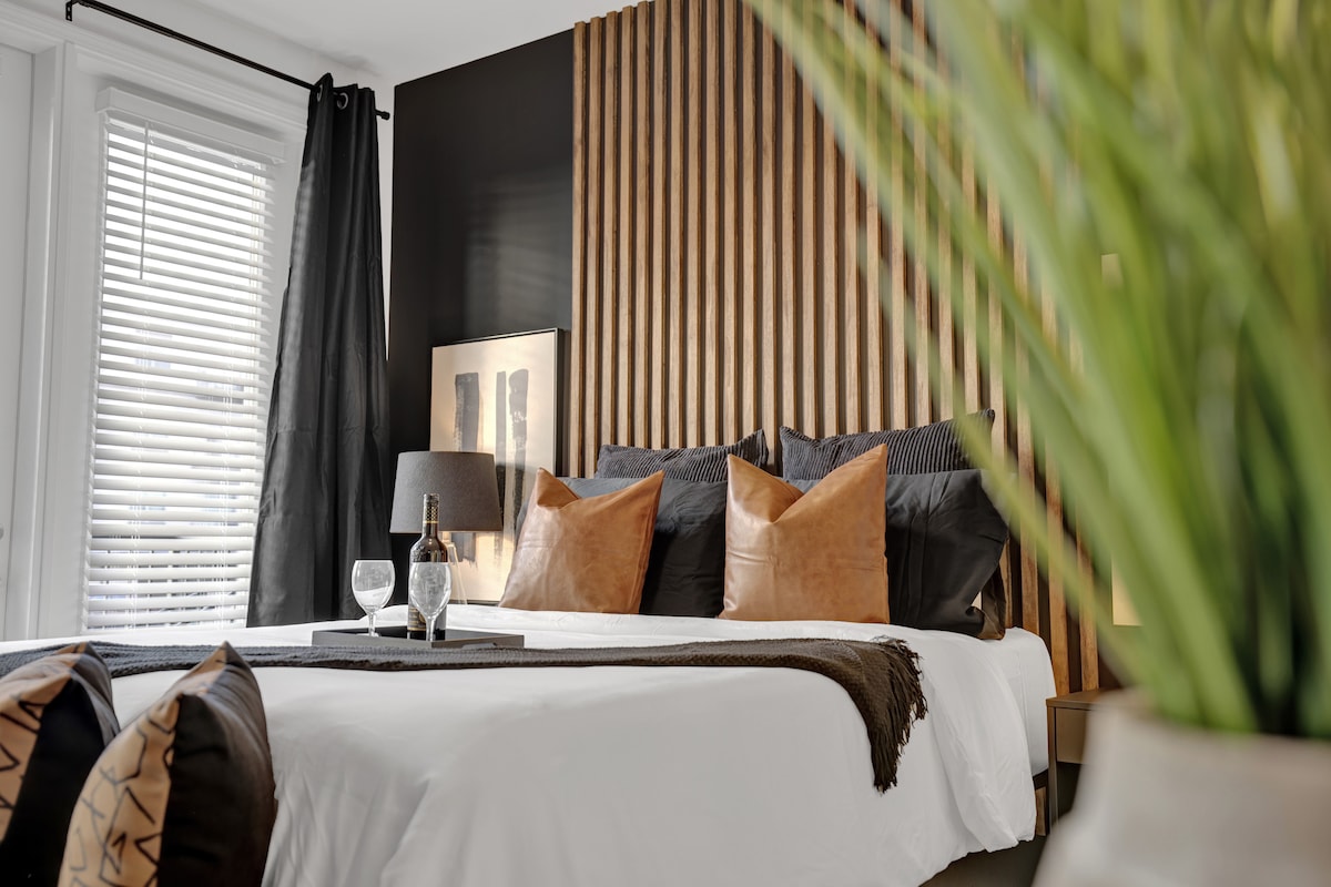 A spacious sleeping area showcases a striking black accent wall paired with vertical wooden paneling. The bed is dressed in crisp white linens, featuring plush pillows in varying tones. A decorative plant can be seen in the foreground, adding a touch of greenery to the scene.
