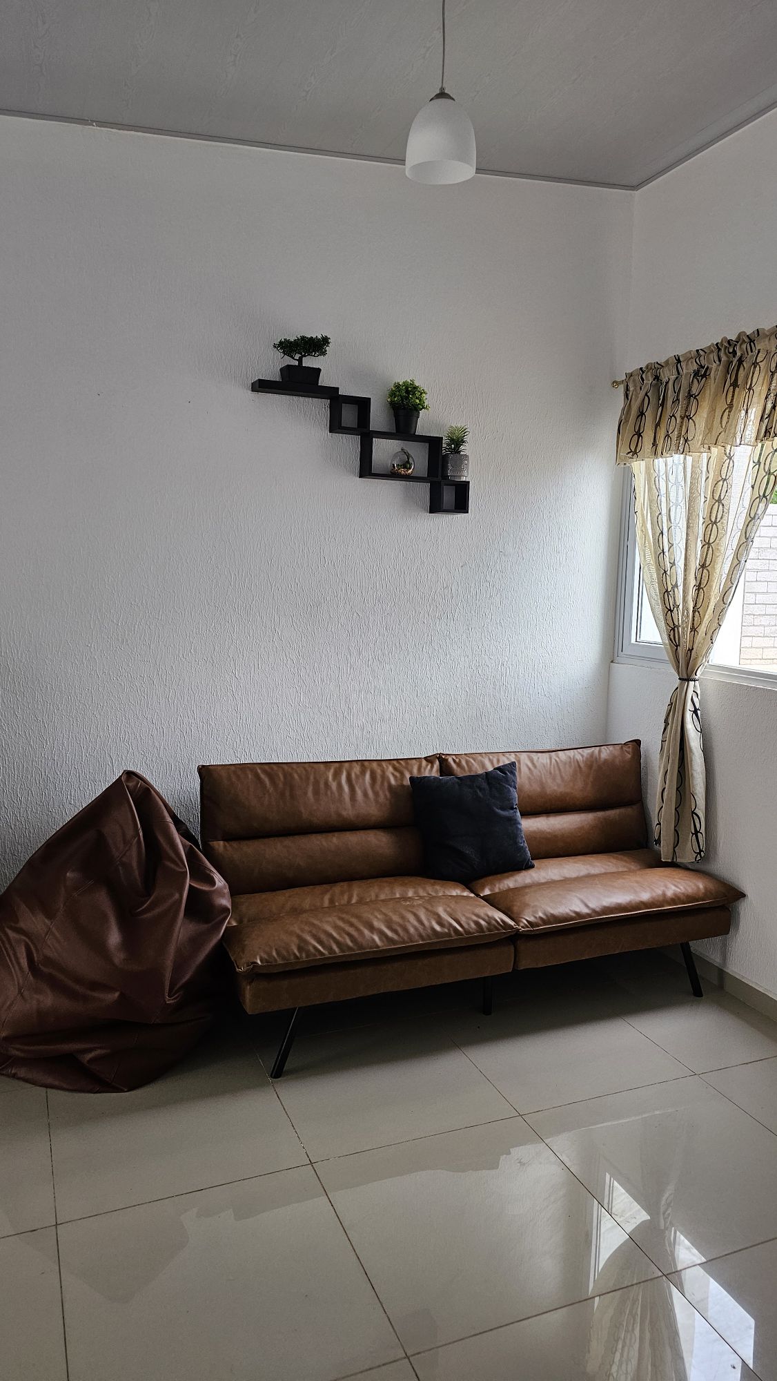 A modern seating area features a brown leather couch accompanied by a decorative black shelf, displaying small plants. Natural light enters through a window adorned with sheer curtains, creating a bright and airy atmosphere. A large bean bag is positioned nearby.
