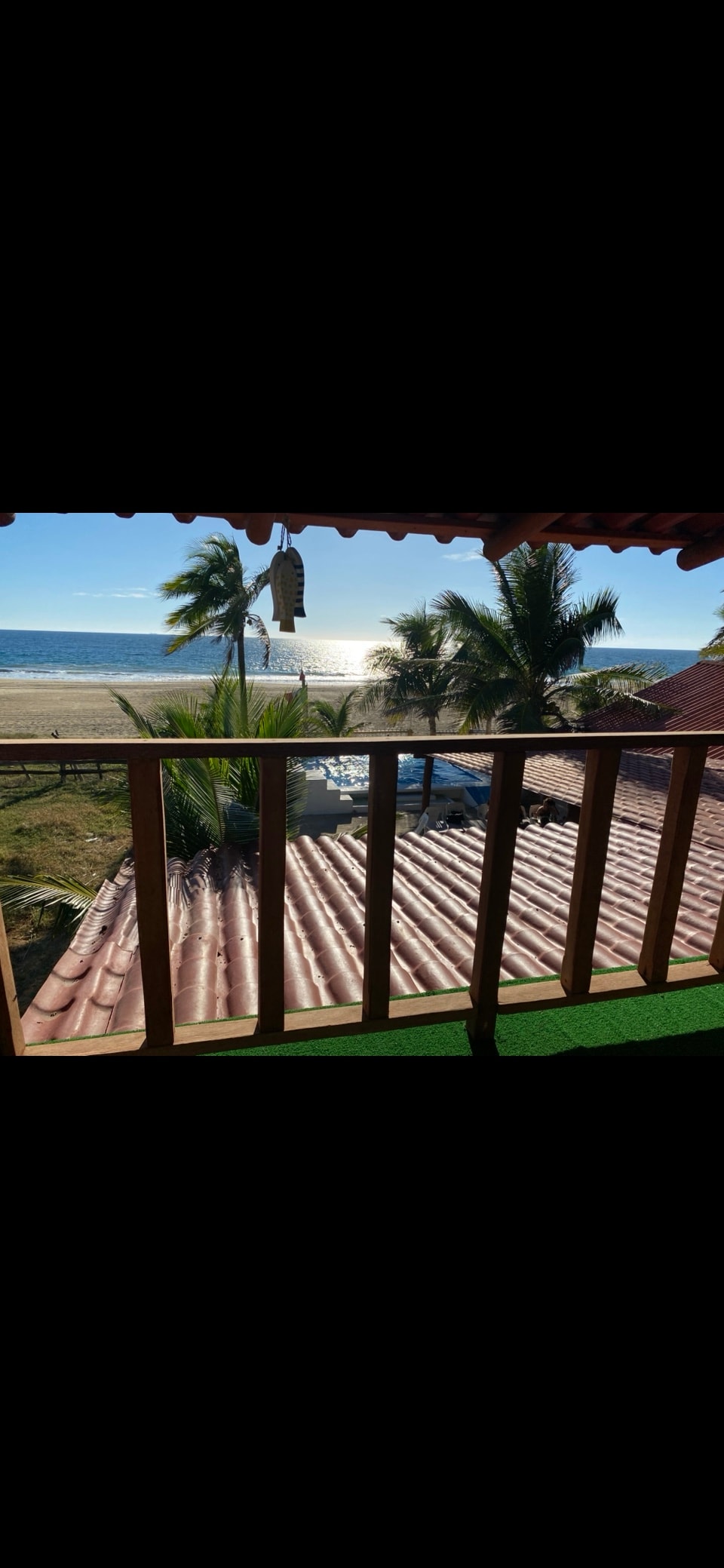 A wooden balcony offers an expansive view of the beach and sea, framed by swaying palm trees. Sunlight reflects off the water, highlighting the sand. A rooftop with terracotta tiles is visible below, creating an inviting coastal atmosphere.