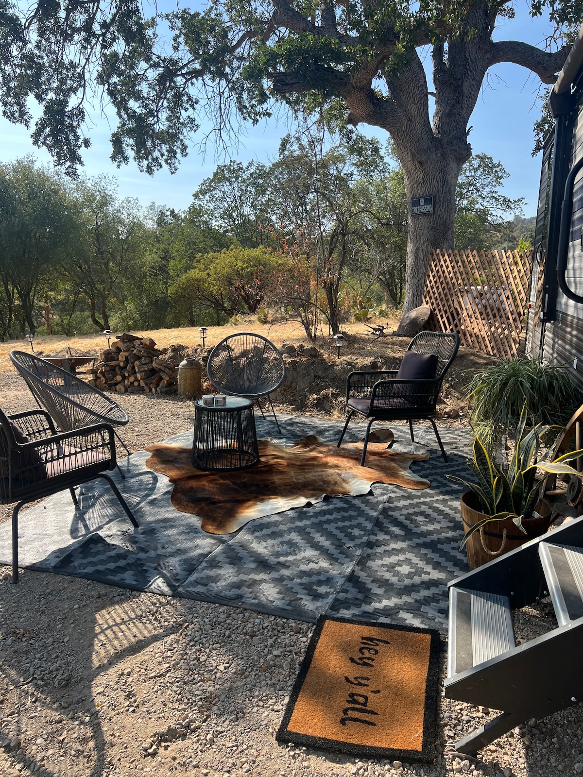 A cozy outdoor seating area is set up on a patterned rug under a shaded tree. Two black chairs and a round table invite relaxation, while a decorative mat greets guests at the entrance. Natural landscaping surrounds the space, enhancing the outdoor experience.