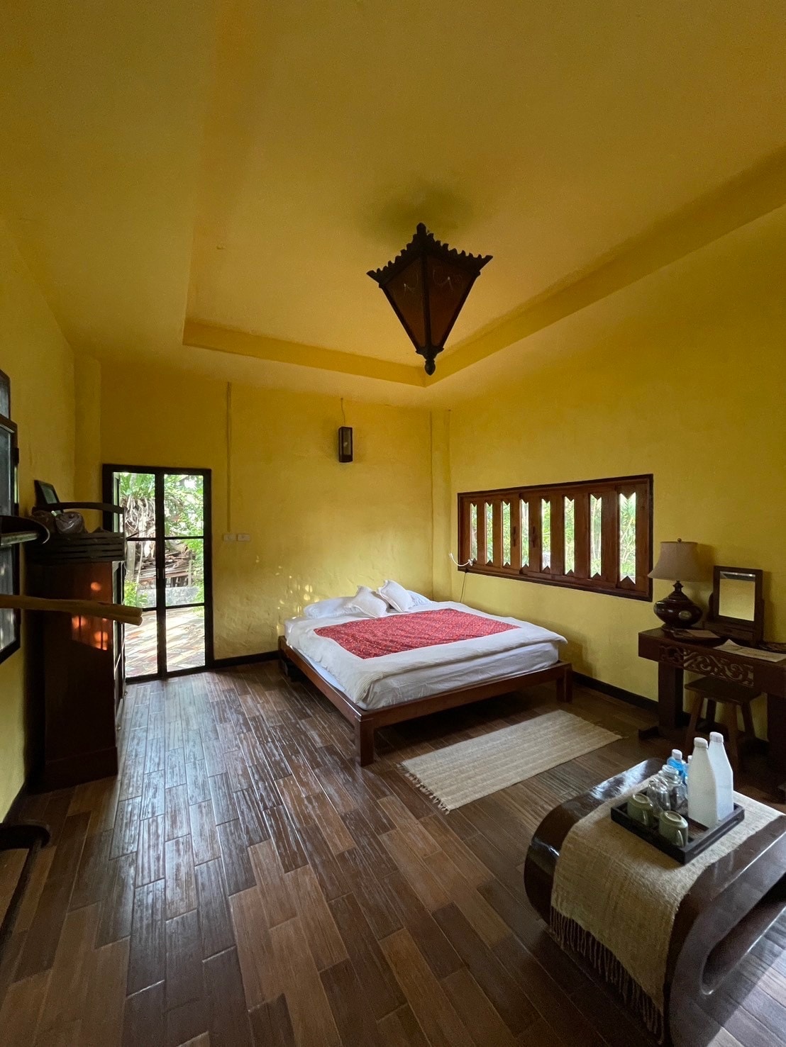 A bright room features yellow walls and a high ceiling. A spacious bed with a red accent blanket is positioned against a wall. Large windows and a glass door offer views of the surrounding nature, while a wooden table displays water bottles and toiletries.