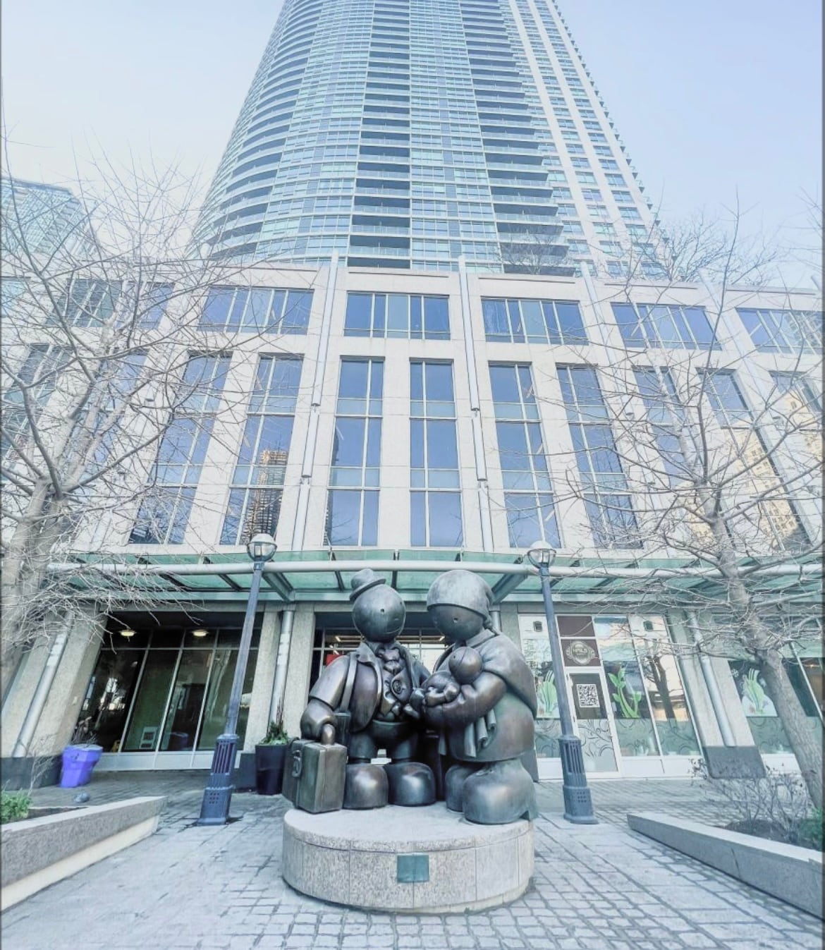 A bronze sculpture depicting two child figures engaged in a playful activity is observed at the base of a towering skyscraper. The building's glass facade reflects the surrounding environment, creating a modern urban atmosphere.