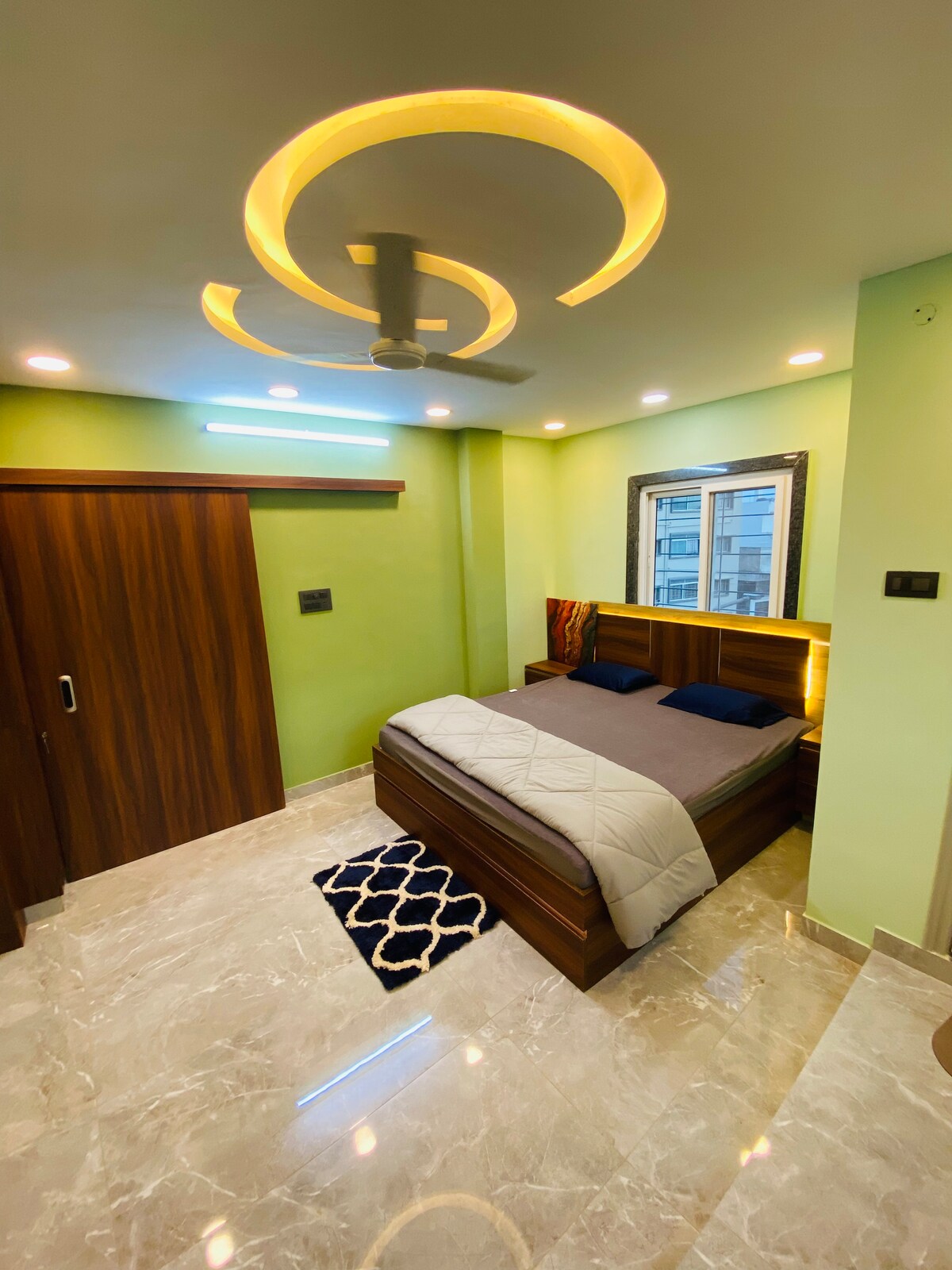 A compact bedroom is presented, featuring a wooden bed with a light-colored blanket and dark pillows. A ceiling fan is visible above, and soft lighting highlights the green walls. A small, patterned rug is positioned by the bed, with a window providing natural light.