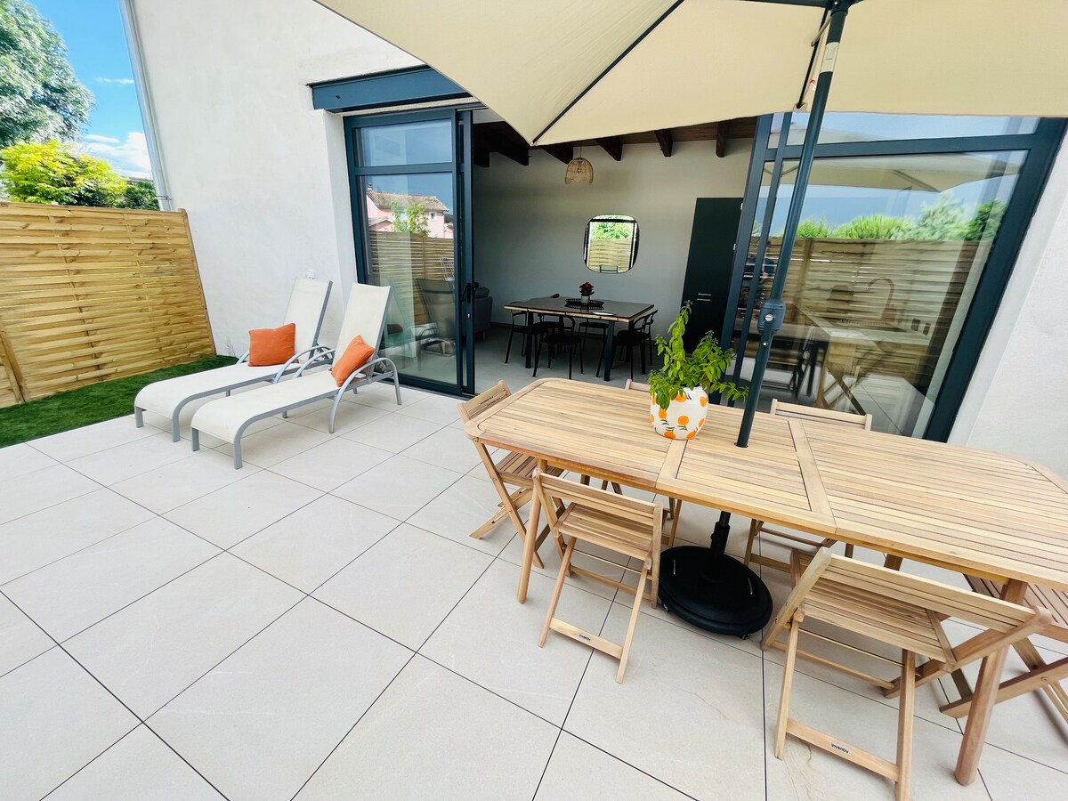 An outdoor terrace features a wooden dining table surrounded by folding chairs and an umbrella. Two sun loungers are positioned nearby, creating a relaxing space. Large glass doors provide access to the interior, allowing natural light to fill the area.