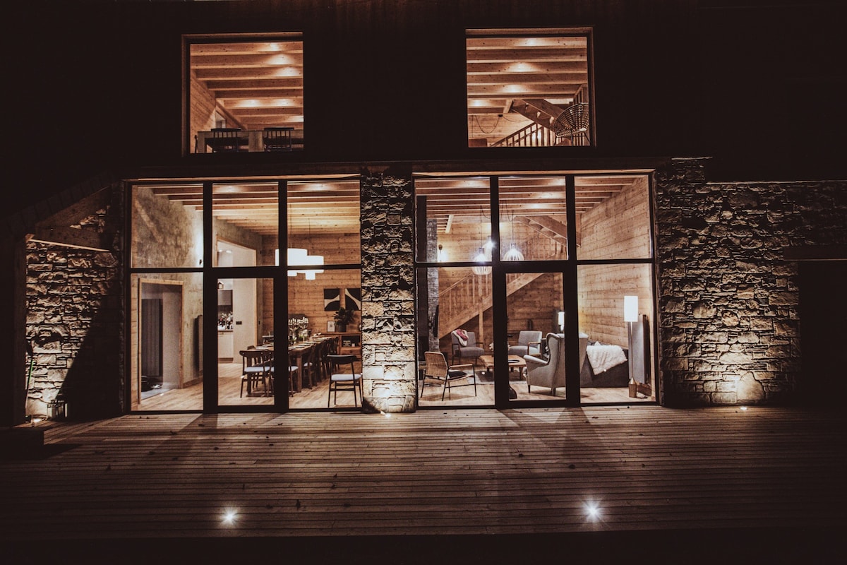 The exterior of the chalet features large glass windows allowing a view of the warm and inviting interiors. Stone walls complement the wood facade. Soft lighting highlights the structure, creating a welcoming ambience for evening relaxation or gatherings.