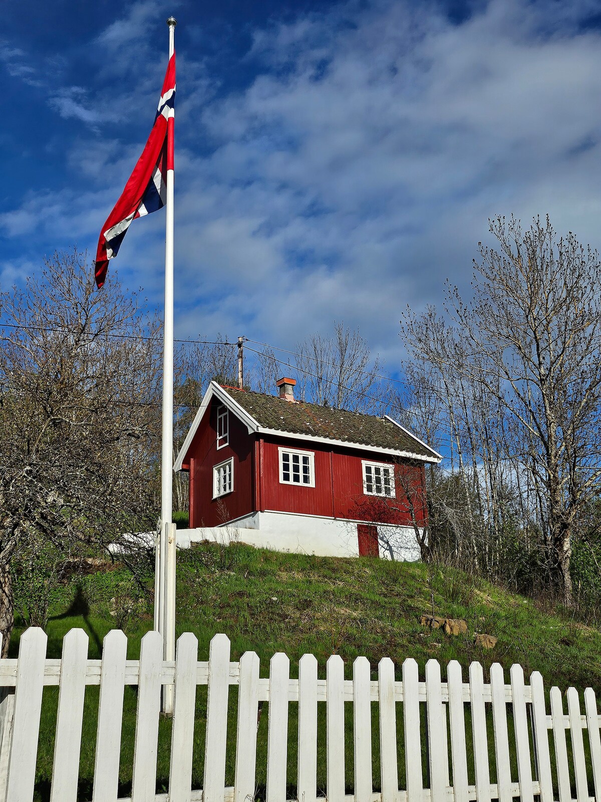 A charming red minihouse perched on a green hill is surrounded by a white picket fence. A Norwegian flag is prominently displayed in front of the house, which features white-framed windows and a sloped roof under a partly cloudy sky.