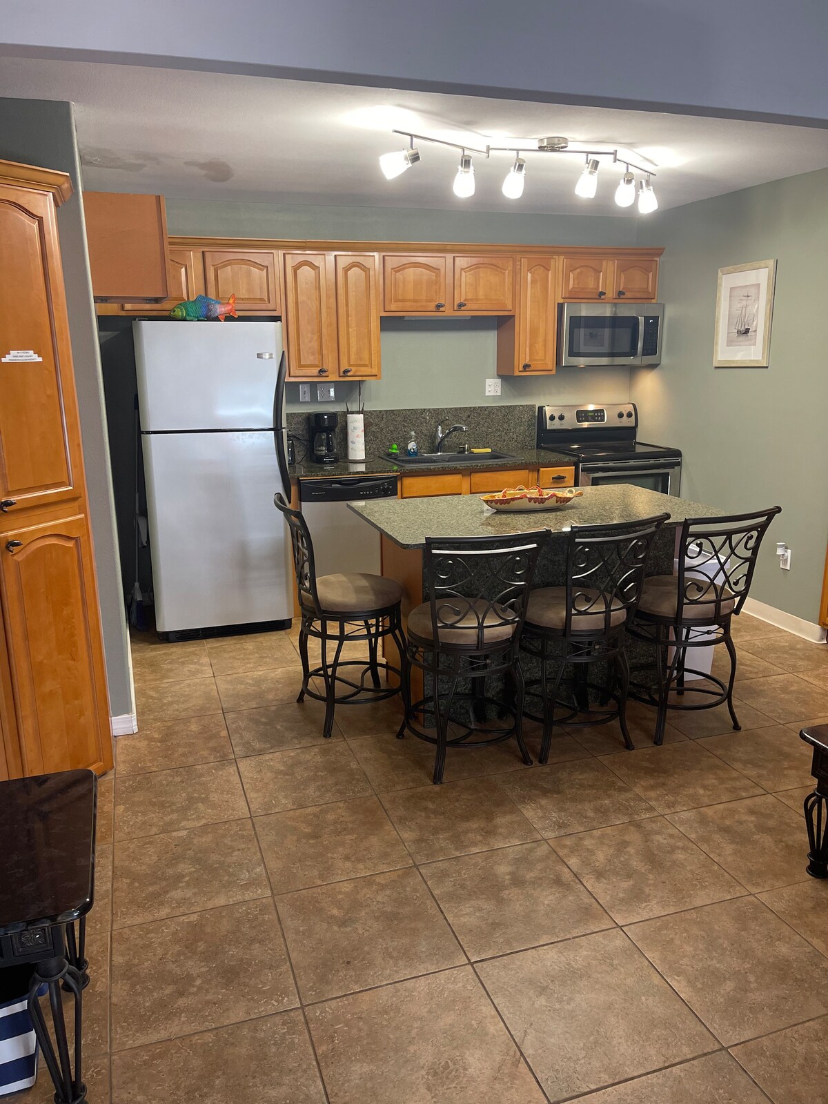 A well-equipped kitchen features wooden cabinetry and granite countertops. Stainless steel appliances, including a refrigerator and microwave, are visible. A spacious island with seating for six provides an area for dining, while soft lighting enhances the warm tones of the room.