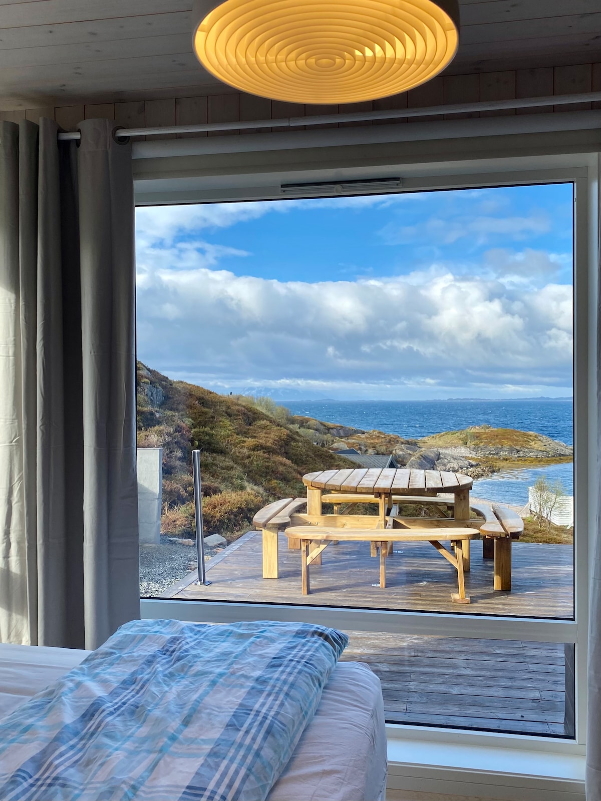 A large window provides a stunning view of the outdoor dining area on a wooden deck, surrounded by natural scenery. The table is set with benches on either side, inviting relaxation while enjoying the sea and mountain landscape beyond.