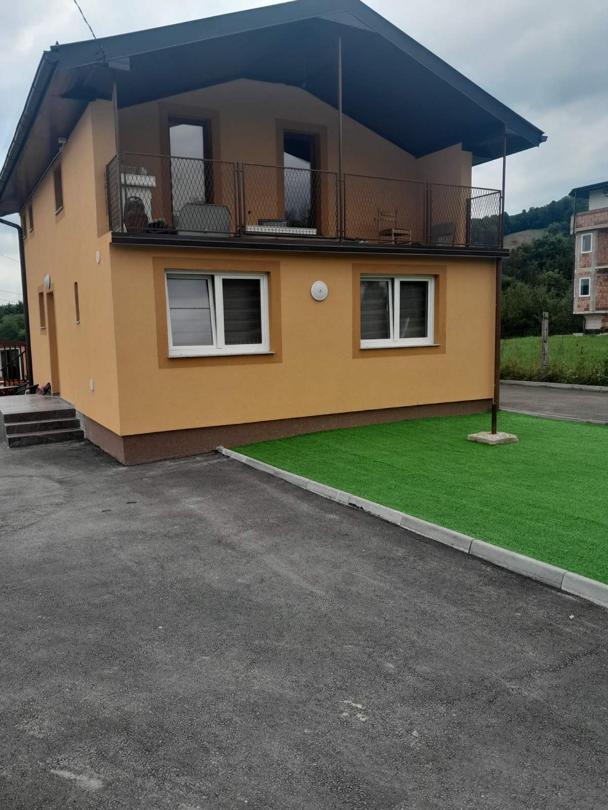 A two-story house is presented with a bright yellow exterior. The first floor features large windows and a small patio area, while a balcony with seating is seen on the upper level. A manicured green lawn complements the paved driveway.