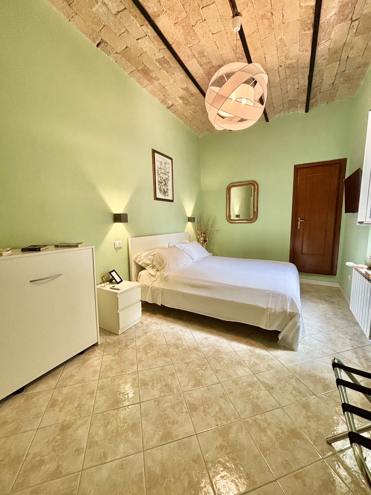 The bedroom features a spacious bed with soft linens, complemented by a decorative mirror and accent light fixtures. A modern light fixture hangs from the ceiling, enhancing the room's simplicity. The tiled floor reflects natural light, creating a serene environment.