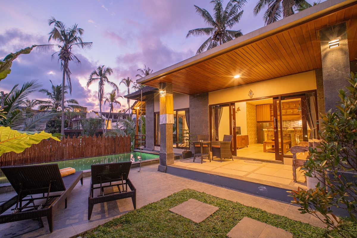 The villa exterior is framed by palm trees under a colorful sky at dusk. A private pool is visible next to a sun bed, and spacious patio doors open to the interior, where a dining area and modern kitchen can be seen.