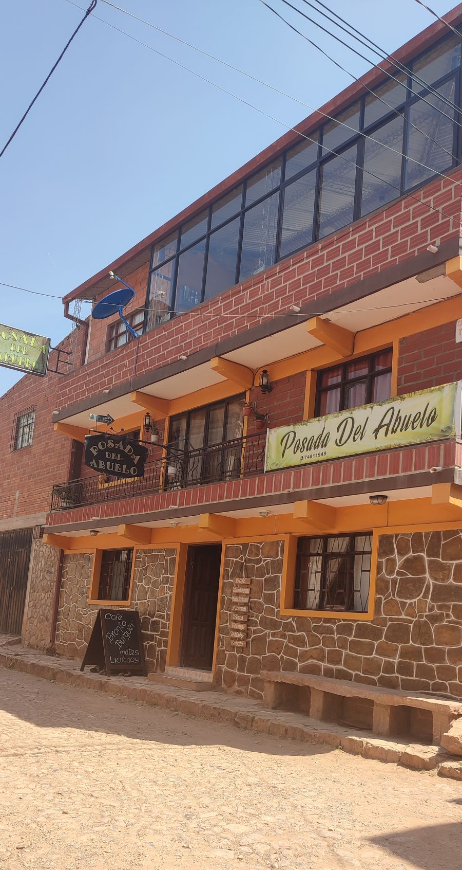 The rustic exterior of a two-story building features a mix of brick and stone materials. Signage for 'Posada Del Abuelo' is visible above the entrance, and balconies with metal railings overlook the street. Natural light brightens the façade, enhancing the welcoming appearance.