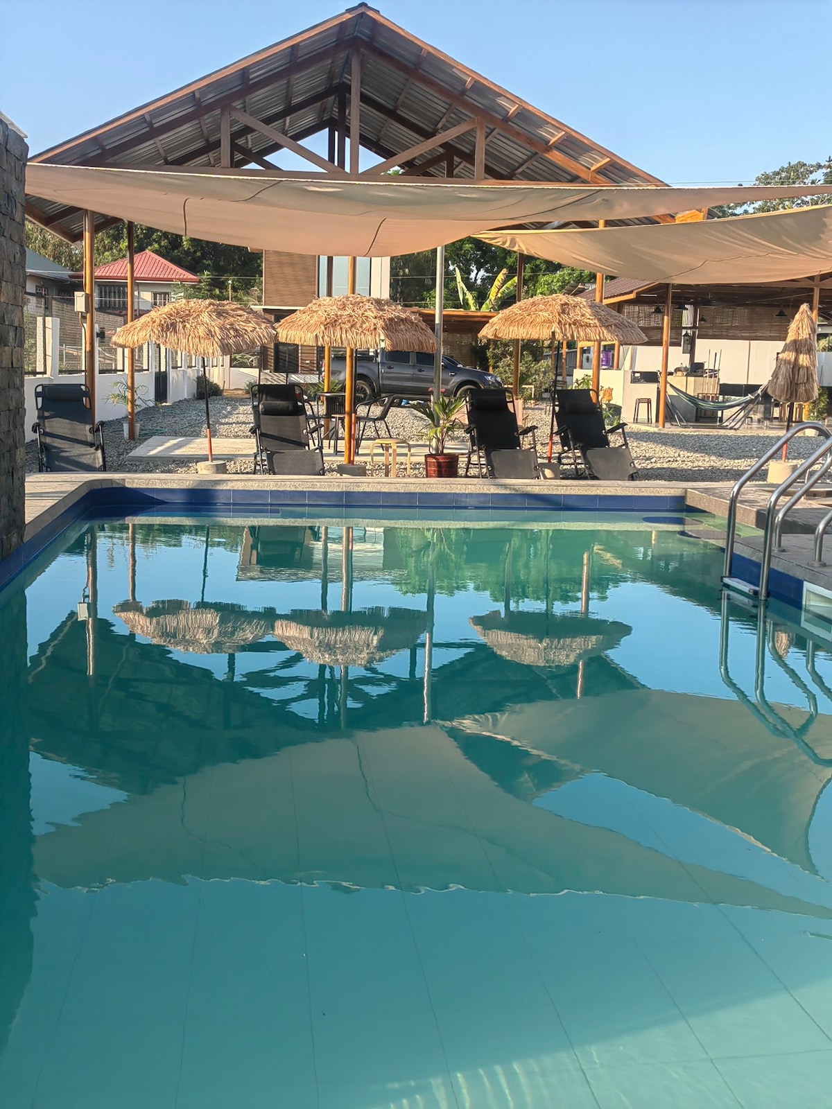 A shared swimming pool glistens under the sun, surrounded by lounge chairs and shaded seating areas with thatched umbrellas. The clear water reflects the serene atmosphere, inviting relaxation and enjoyment.