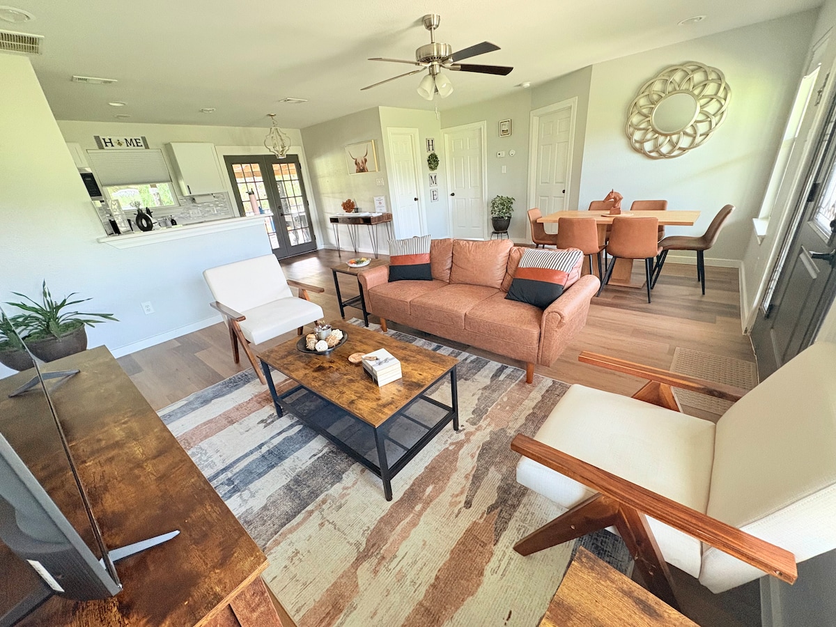 The open living area features a comfortable sofa, two armchairs, and a wooden coffee table. Natural light fills the space through large windows. A dining area with modern chairs is visible in the background, alongside a kitchen with an inviting design.