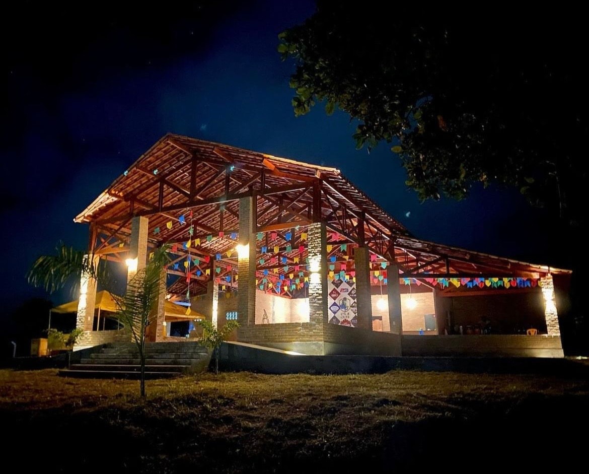 A spacious open structure is seen at night, featuring a colorful canopy of decorations and bright lighting. The design emphasizes natural materials and is surrounded by lush greenery, creating a welcoming ambiance for gatherings.