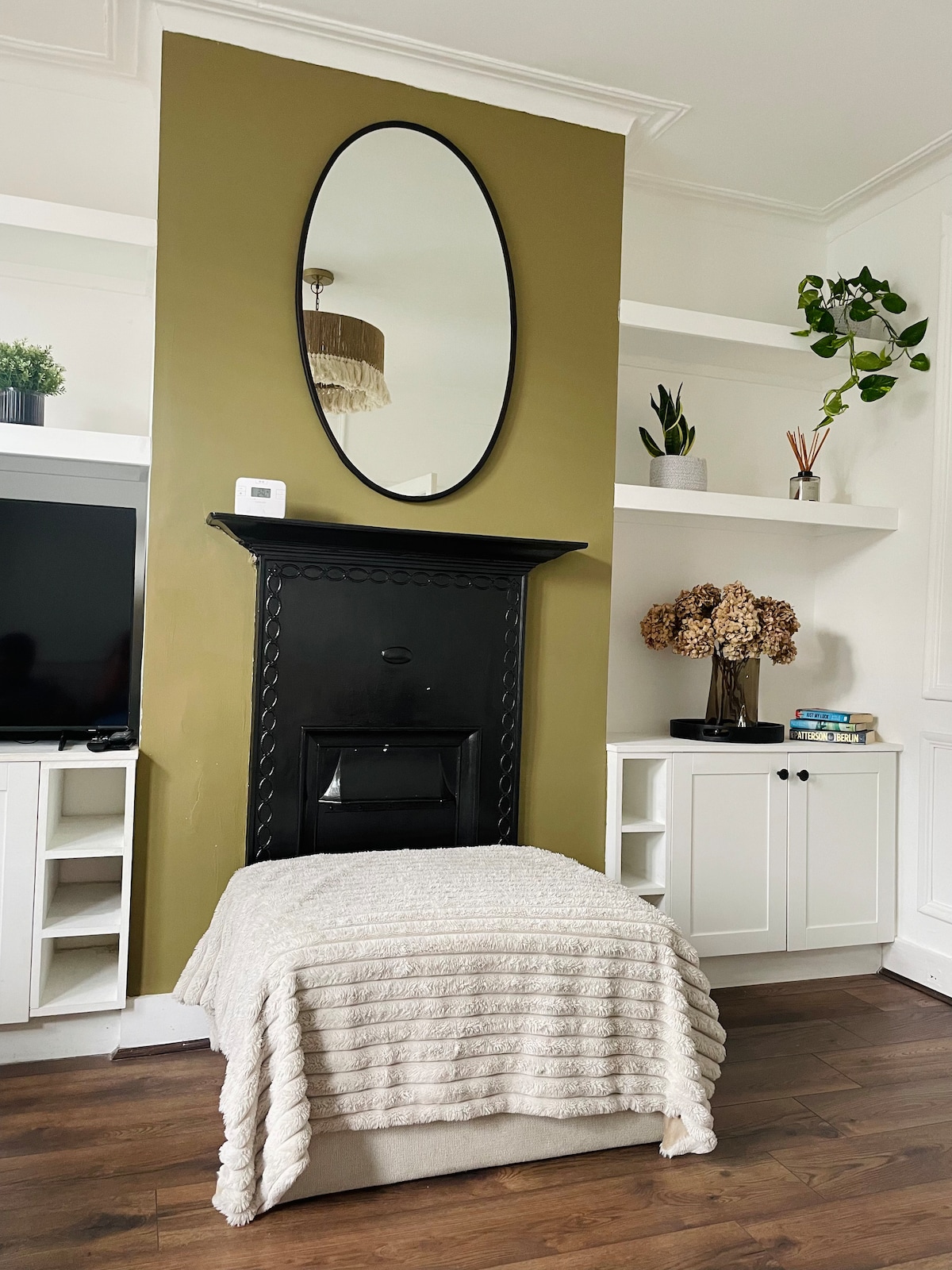 A welcoming living space features a tall mirror above a black fireplace, surrounded by a soothing olive-green wall. Shelves display decorative plants and books. A soft throw blanket is draped over a cozy ottoman, offering a touch of comfort to the room.