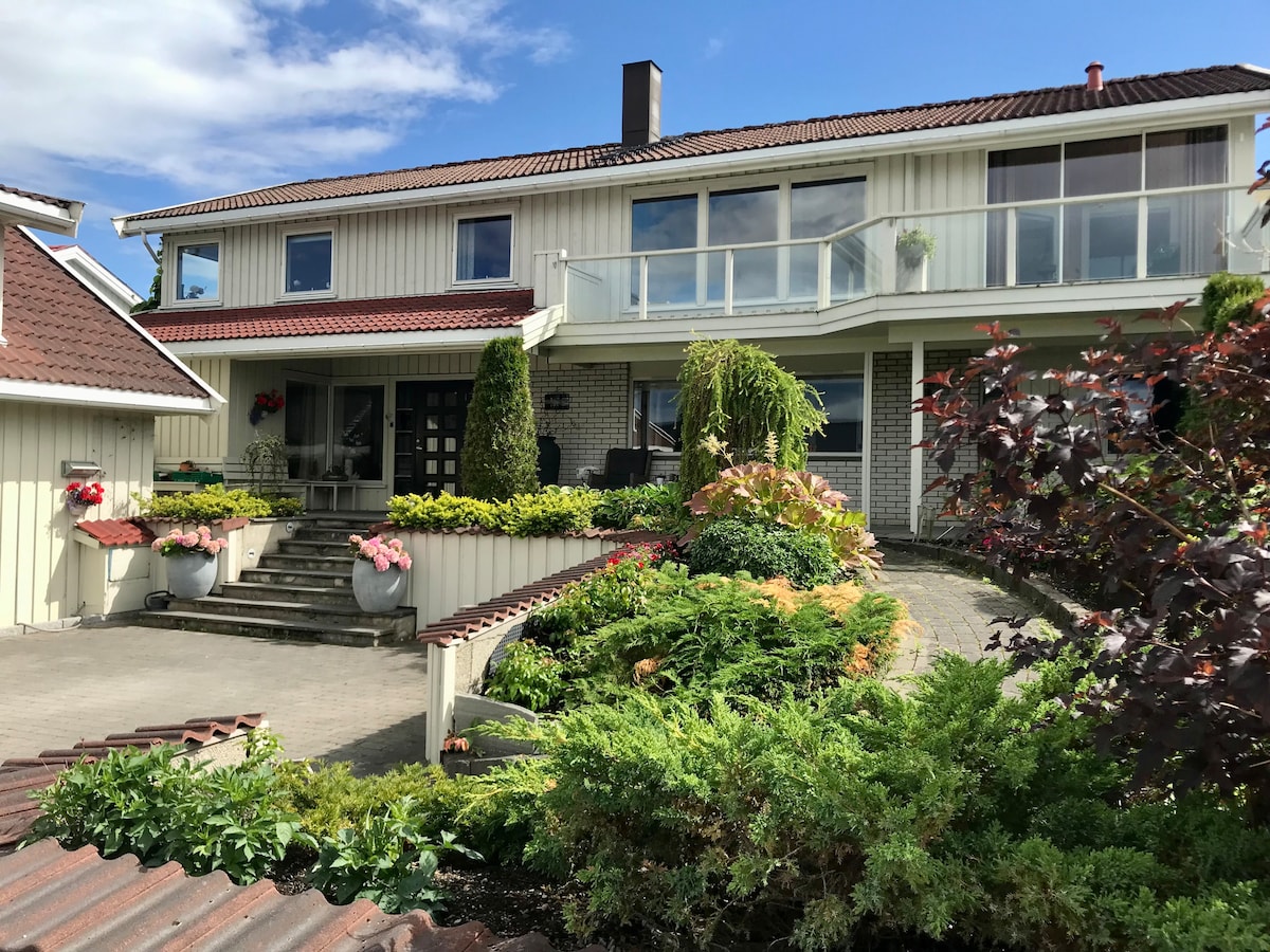 The exterior of a well-maintained house is shown, featuring multiple levels and large windows that provide natural light. Surrounding gardens with vibrant flowers add color. A stone pathway leads to the entrance, while greenery offers a lush backdrop, contributing to a serene outdoor environment.