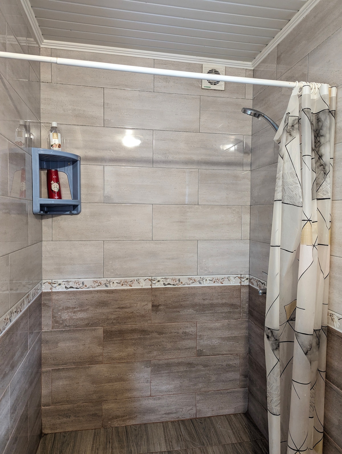 A modern shower space features tiled walls in neutral tones, with a showerhead positioned above. A decorative border runs along the midsection, and a shower curtain offers privacy. A wall-mounted shelf holds personal care items, providing convenient storage.