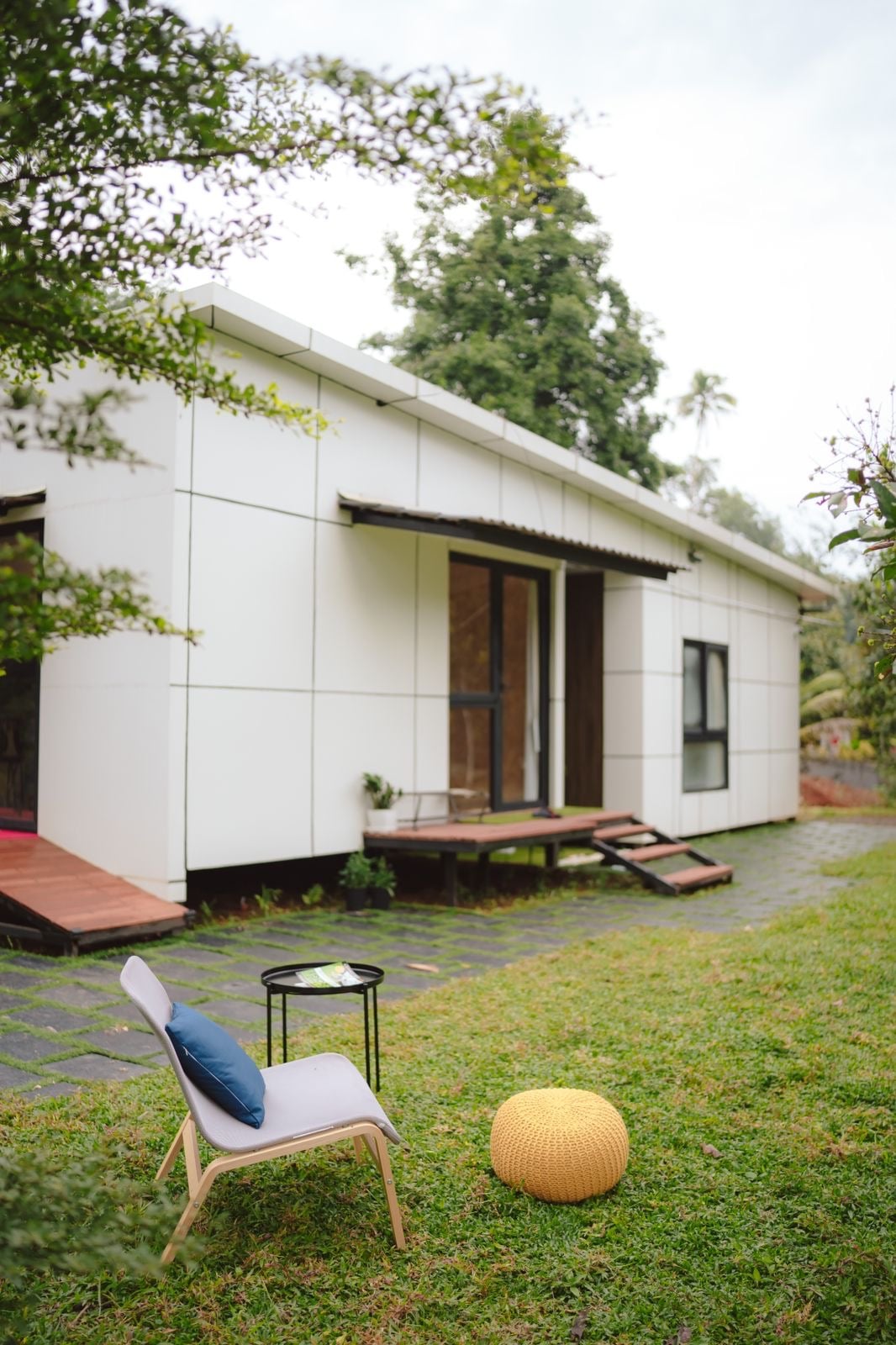 A minimalistic exterior of a villa is shown, featuring two entrances with wooden steps leading up to them. A small seating area with a chair and a pouf sits on a grassy lawn, with lush greenery surrounding the space.
