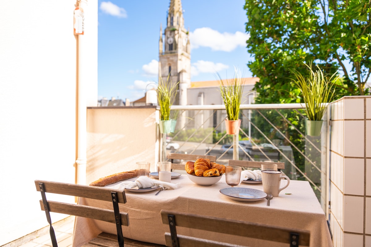 An outdoor dining area is set with a table covered in a neutral cloth, featuring plates, mugs, and a basket of fresh croissants. Green plants are placed in wall-mounted pots, with a backdrop of a church tower and blue skies.