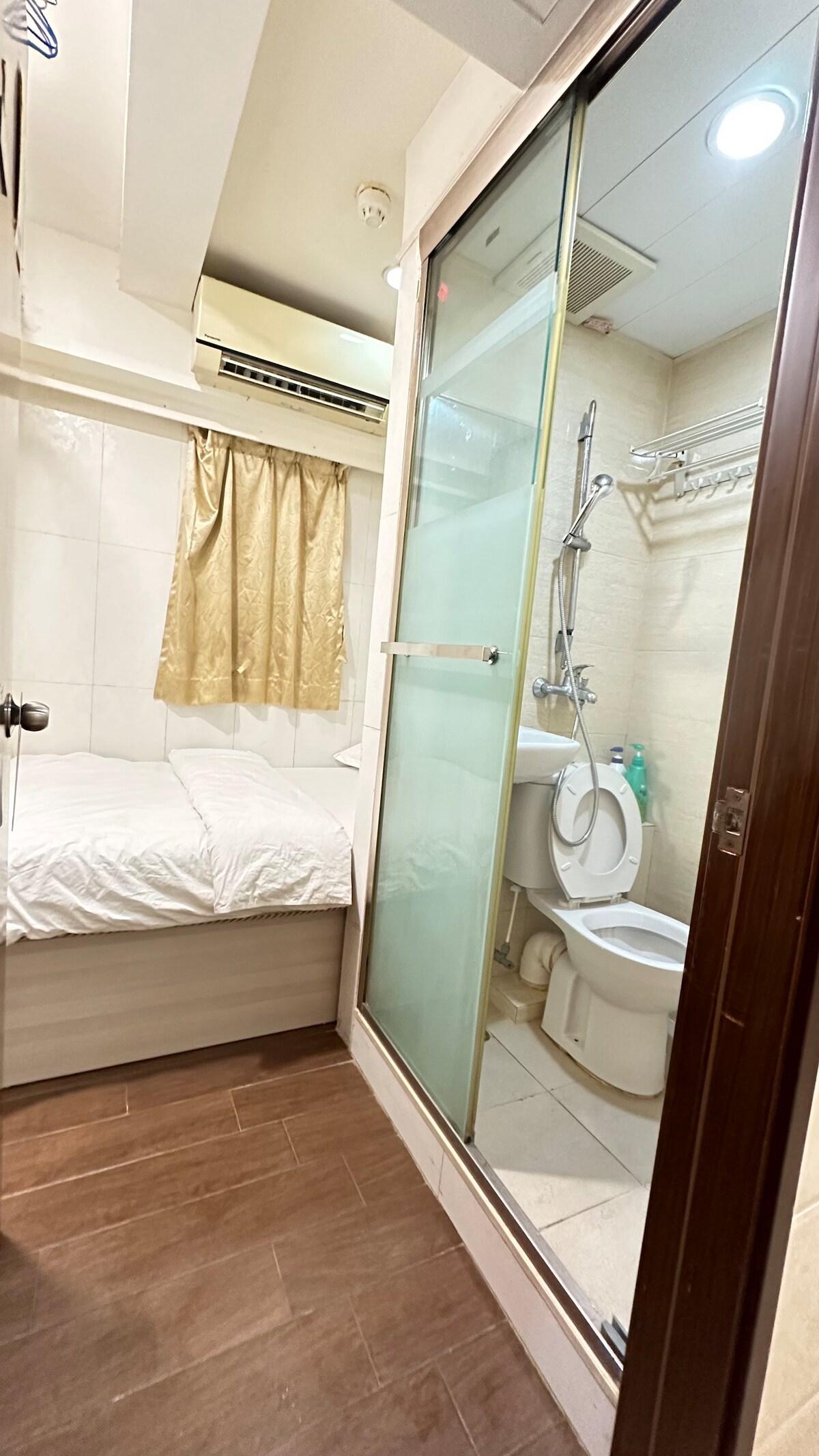 A compact bathroom is positioned adjacent to a sleeping area. The space features a glass shower enclosure and a toilet. Natural light enters through a curtain. The sleeping area is equipped with a bed dressed in white linens, contributing to a clean ambiance.