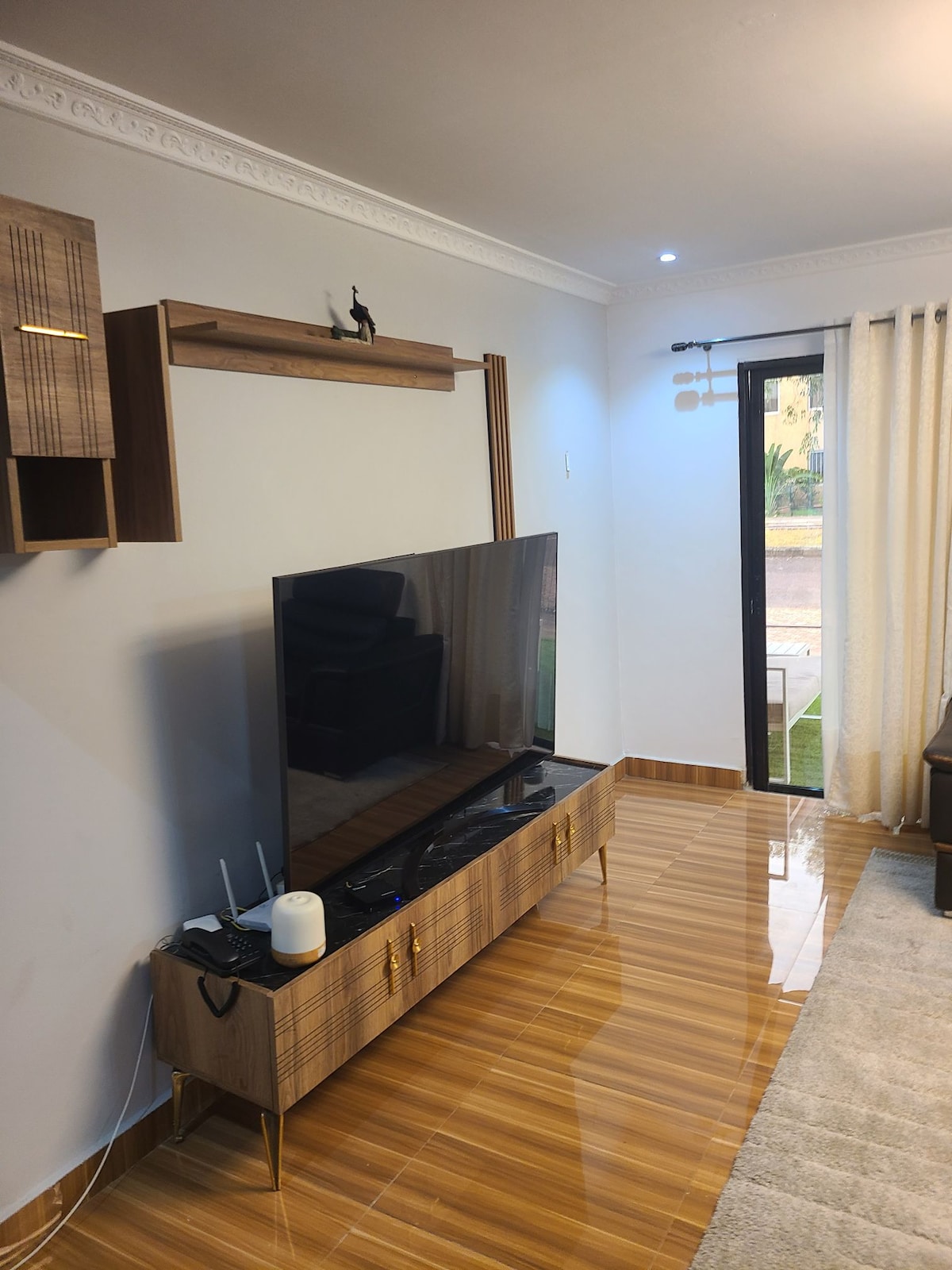 A modern living space features a sleek flat-screen television mounted above a stylish, low-profile media console. The floor shines with polished tiles, and soft light is provided by wall sconces. Light filters through sheer curtains, illuminating the room's contemporary design.