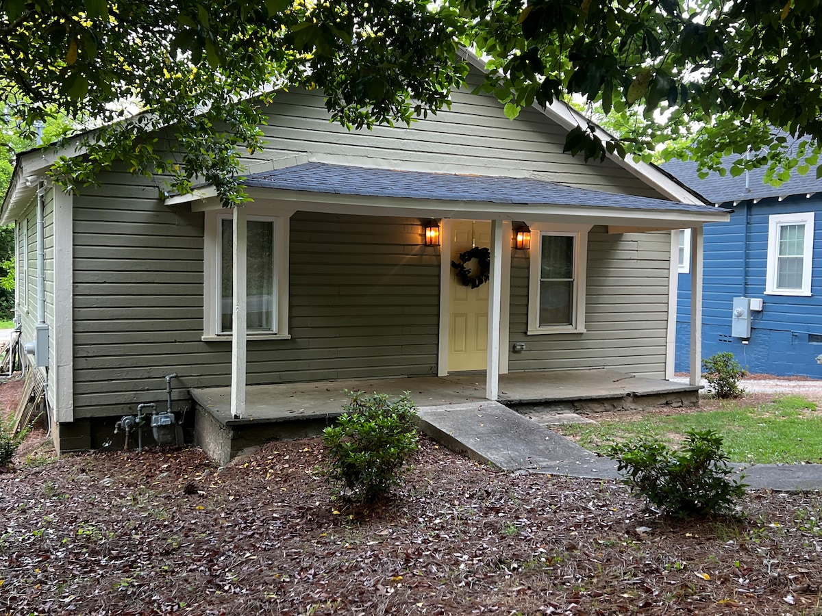 A single-story cottage features a welcoming porch with two light fixtures and a decorative wreath on the door. The exterior showcases earthy green siding, complemented by surrounding shrubs and a gravel pathway. Trees provide shade in the front yard, enhancing the cottage's curb appeal.