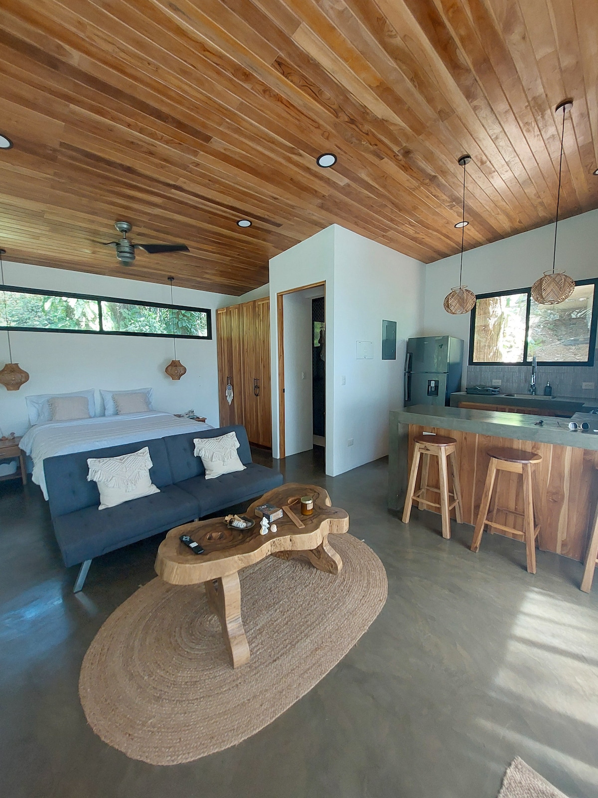 A studio space features a queen bed and a fold-down couch, complemented by natural wood accents on the ceiling. A kitchen with modern appliances and bar stools is visible, while a round woven rug anchors the seating area. Large windows allow natural light to fill the room.