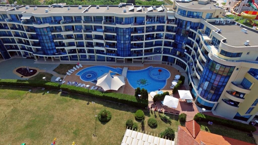 An aerial view showcases a curved building surrounded by lush greenery. A serene swimming pool with shaded areas and lounge chairs is visible in the foreground, complemented by various seating arrangements under fabric canopies. The modern architecture reflects sunlight through large glass windows.