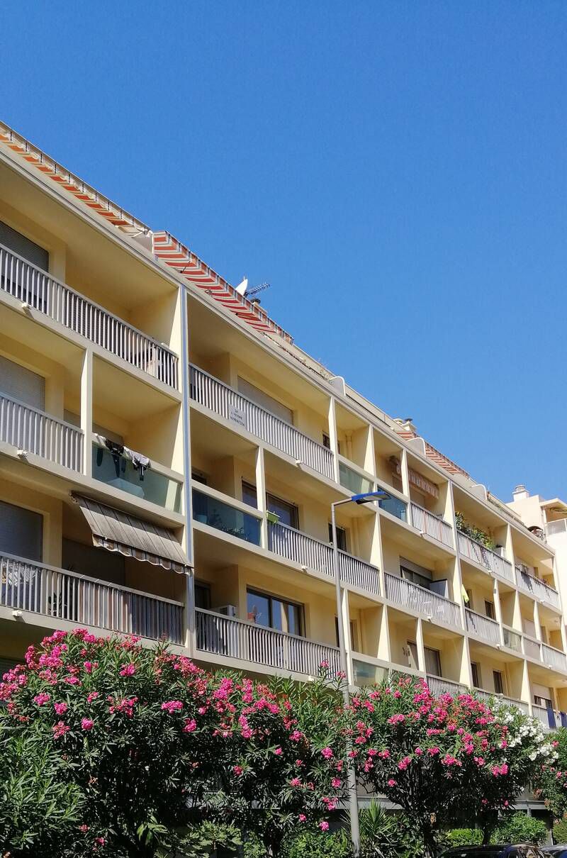 A multi-story residential building is depicted against a clear blue sky. Balconies with railings extend from each level, some adorned with plants and clothes. The vibrant green foliage of nearby blooming shrubs adds color to the foreground, enhancing the building's welcoming appearance.