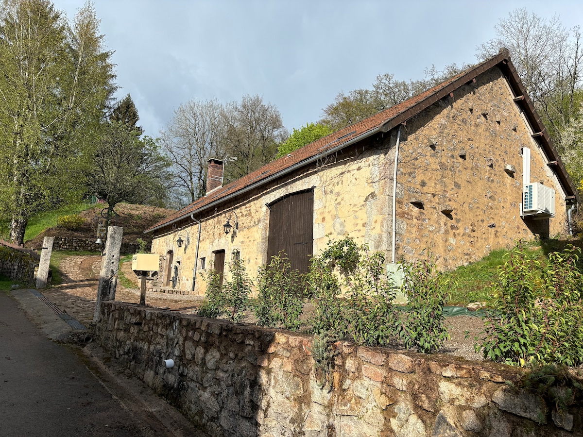 The rustic stone exterior of the longère is presented, featuring a sloped roof and wooden double doors. Surrounding greenery and a gravel pathway enhance the entrance. A stone wall borders the property, while trees and shrubbery provide a natural backdrop.
