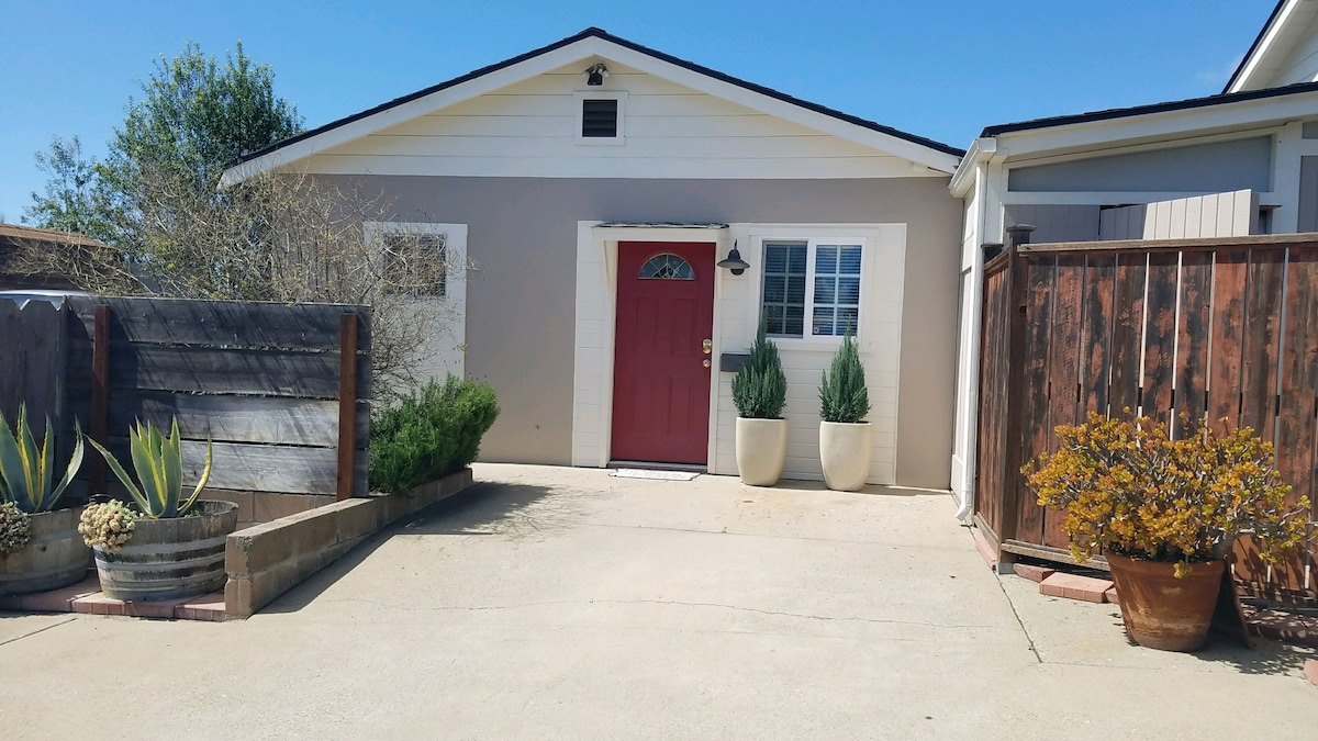 A welcoming exterior features a bright red front door, complemented by two large planters filled with greenery. The entrance is framed by a well-manicured concrete driveway, with natural elements like a wooden fence and decorative pots enhancing the inviting atmosphere.