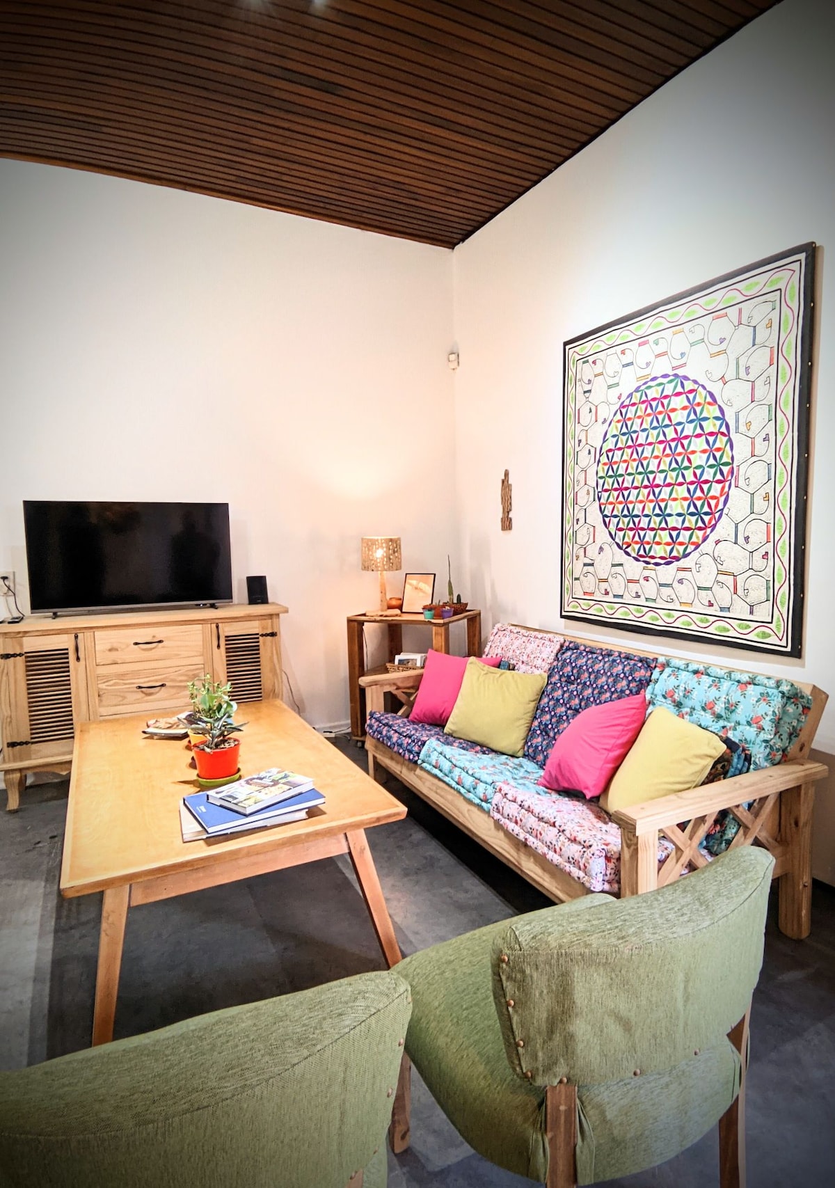 The living area features a wooden coffee table positioned at the center, surrounded by a colorful couch with patterned cushions and two accent chairs. A flat-screen television is mounted on the wall, and a vibrant piece of artwork hangs above the couch, adding character.