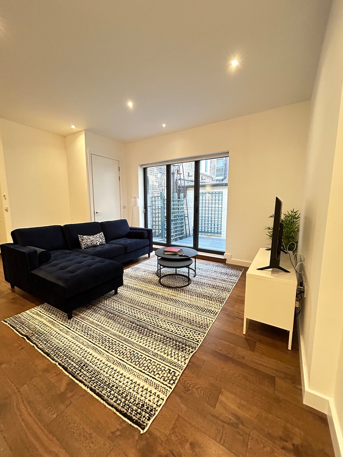 The living area features a navy blue sectional sofa positioned beside a small round coffee table. A patterned rug covers the wooden floor, while sliding glass doors open to a private outdoor space. A flat-screen TV is mounted on the opposite wall.
