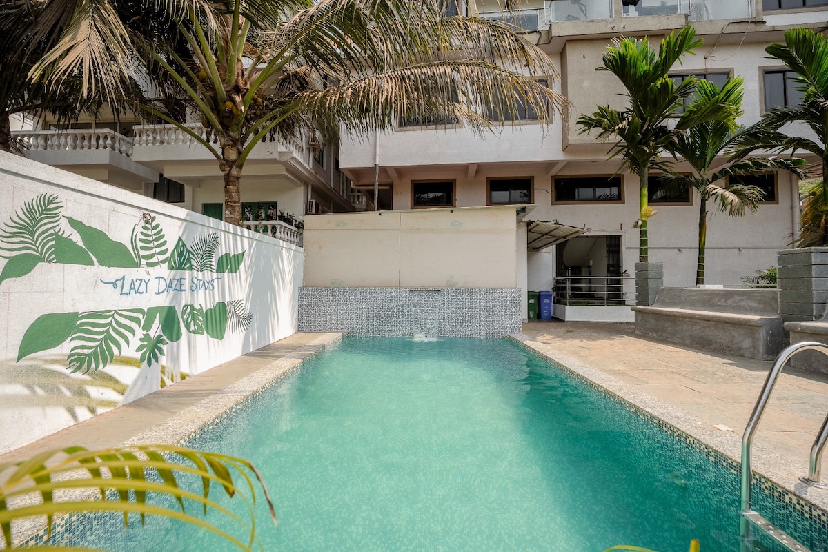 A serene swimming pool is shown, with clear turquoise water reflecting sunlight. Lush palm trees frame the area, and there is a decorative mural featuring green leaves on the white wall adjacent to the pool. The surrounding space appears spacious and inviting.
