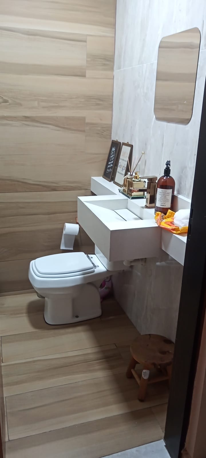 A modern bathroom features warm-toned wooden paneling and beige marble walls. A wall-mounted sink with a storage space underneath is positioned beside a white toilet. A small stool and decorative items are present, adding a touch of design, while a mirror reflects the area.