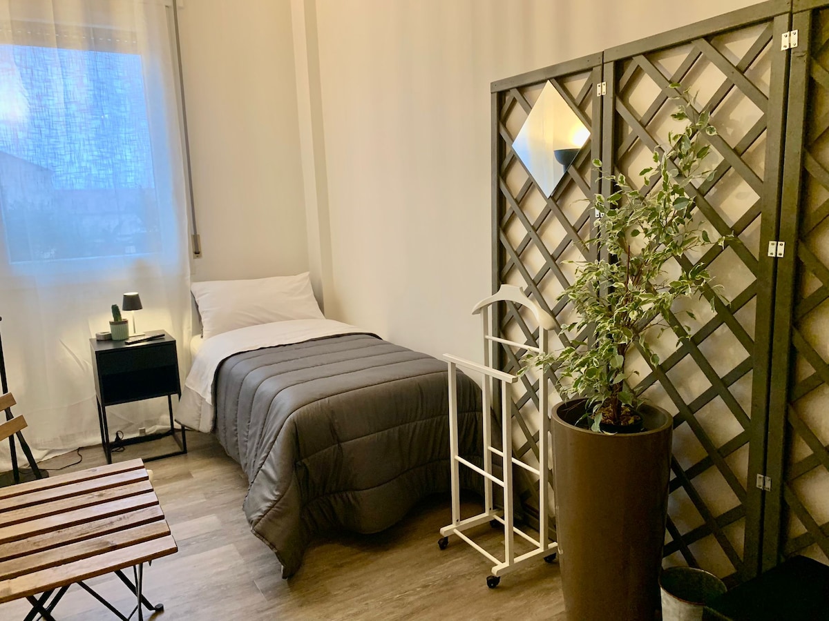 A single bed is arranged with a gray blanket, complemented by a small nightstand featuring a lamp. A decorative lattice screen separates the space, while a potted plant adds a touch of greenery. A wooden table and chair provide additional seating options.