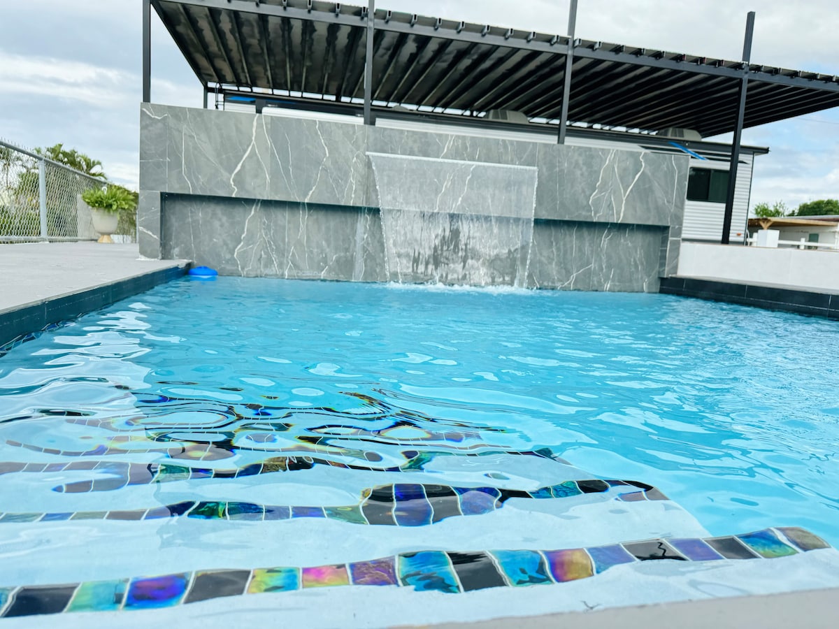 A clean pool area is shown with clear blue water, featuring a modern waterfall cascading over a polished stone wall. Colorful tiles adorn the pool's edge, complementing the serene environment while providing an inviting place for relaxation.