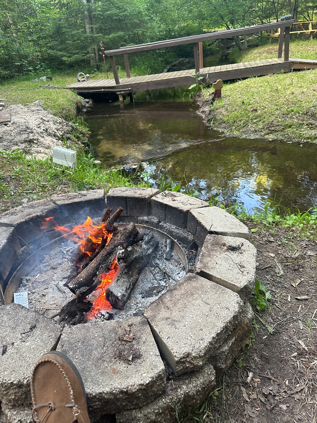 A stone fire pit is surrounded by grass, with a small fire burning brightly at its center. A wooden bridge crosses over a gently flowing creek in the background, surrounded by lush greenery. The tranquil setting invites outdoor relaxation.