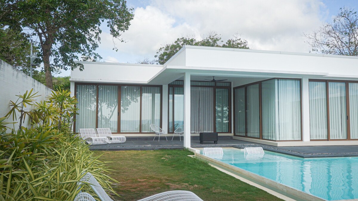 A contemporary villa showcases large glass sliding doors, providing abundant natural light. The sleek design features a private pool surrounded by a manicured lawn and sun loungers. Lush greenery lines the property, inviting relaxation in a tranquil outdoor setting.