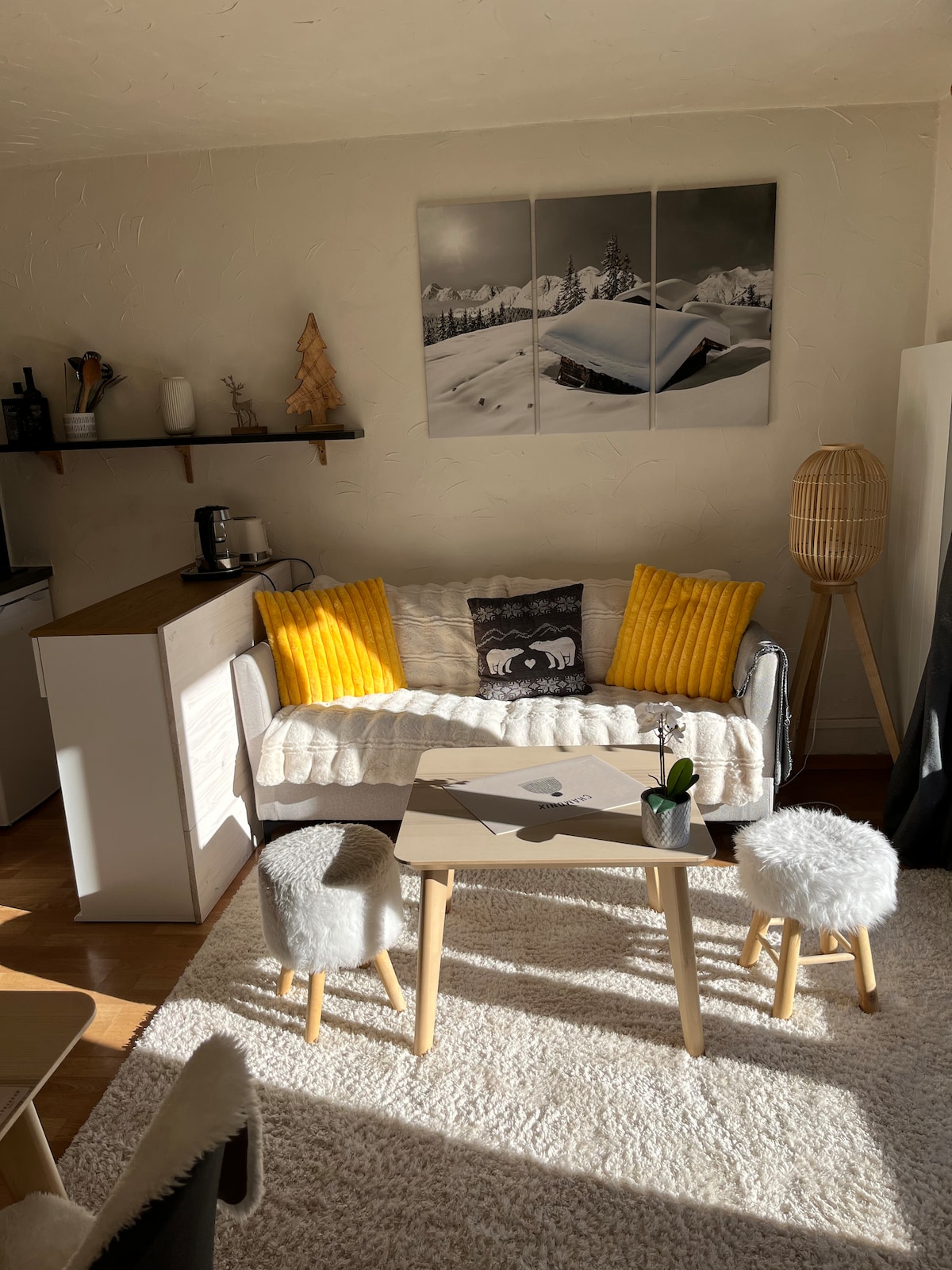 A cozy living area features a light-colored sofa adorned with yellow cushions, set against a backdrop of scenic mountain artwork. A wooden coffee table is placed in front, accompanied by two soft, furry stools. Natural light fills the space, highlighting the warm tones of the decor.