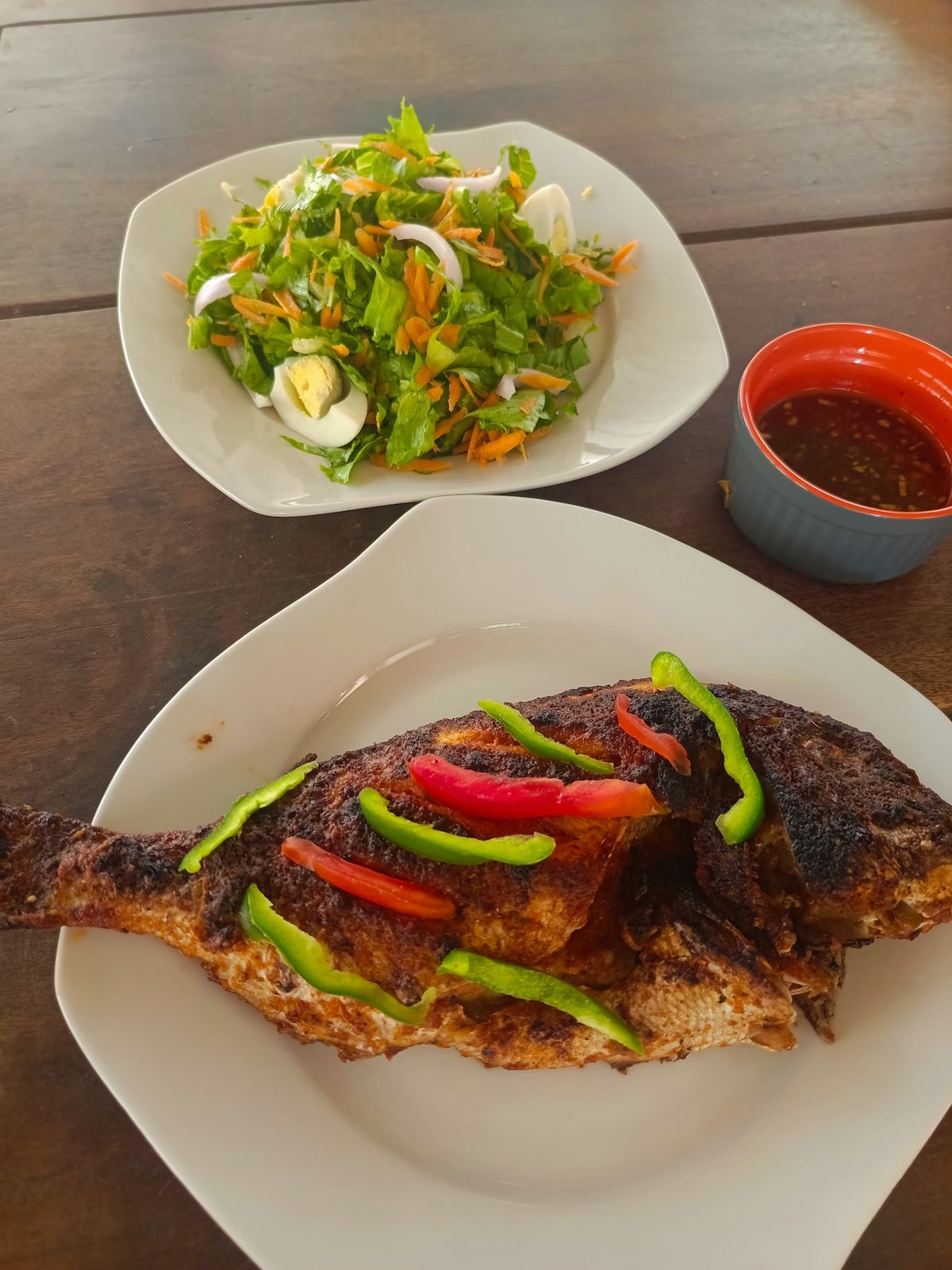 A plated meal is presented featuring a whole fish garnished with colorful strips of red and green peppers. Beside it, a fresh salad is served, consisting of mixed greens, shredded carrots, and halved boiled eggs. A small bowl of dipping sauce accompanies the dishes.