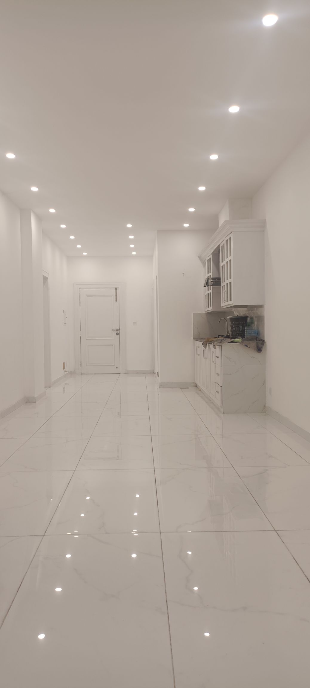 An elongated hallway features glossy white tiles reflecting overhead lighting. The white walls provide a clean backdrop, while a white kitchen cabinet is seen along one side, accompanied by minimalist decor. A door is located at the end of the hallway, enhancing the spacious feel.