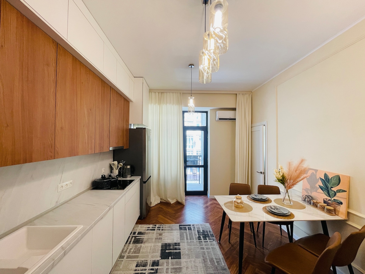The kitchen area features modern cabinetry with a sleek design and a fully equipped cooking space. A dining table is set for two, complemented by stylish chairs. Natural light filters through a large window, illuminating the warm wood flooring and tasteful decor.